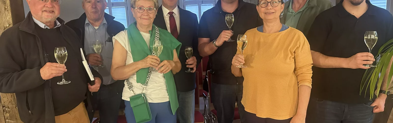 Gruppe von acht Personen bei einer Veranstaltung in einem historischen Raum mit Holzbalken. Die Teilnehmenden halten Sektgläser in der Hand und schauen in die Kamera.