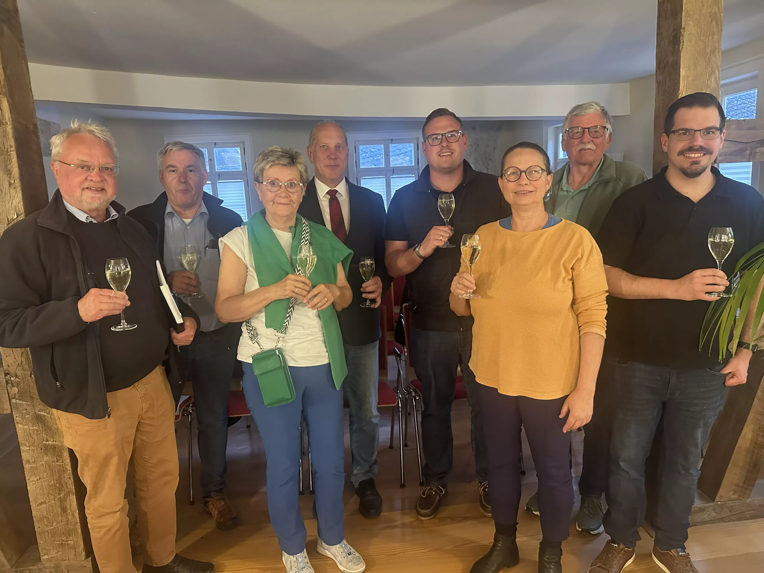 Gruppenfoto bei einem gemeinsamen Empfang Gruppe von acht Personen bei einer Veranstaltung in einem historischen Raum mit Holzbalken. Die Teilnehmenden halten Sektgläser in der Hand und schauen in die Kamera.